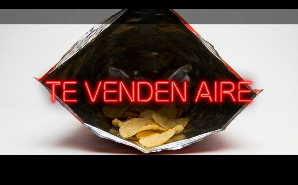 Fotografía de producto cinemática en formato 16:10 con franja negra inferior. Una bolsa de patatas fritas cortada transversalmente, mostrando que el 80% del interior es aire vacío y solo hay unas pocas patatas tristes en el fondo. Iluminación de contraste alto. Sobre la imagen, texto en neón rojo: "TE VENDEN AIRE".
